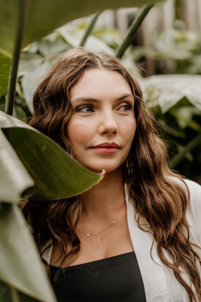 A woman with a soft, reflective expression stand among palm leaves, gazing past the camera with quiet strength.  