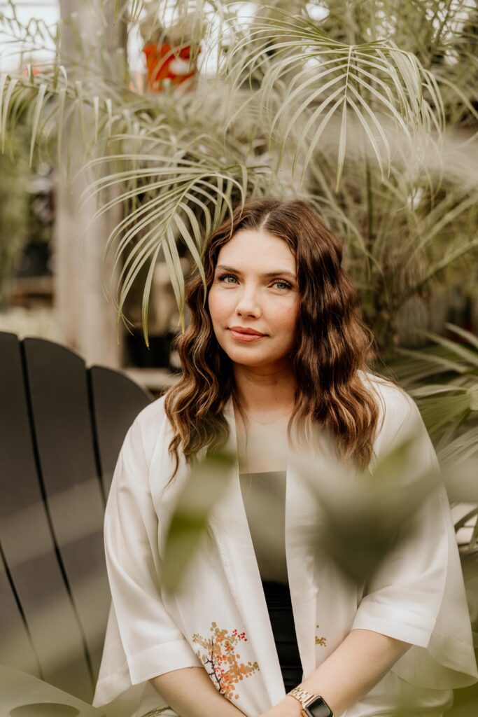 A woman sits calmly in a garden among palm leaves looking reflectively at the camera.