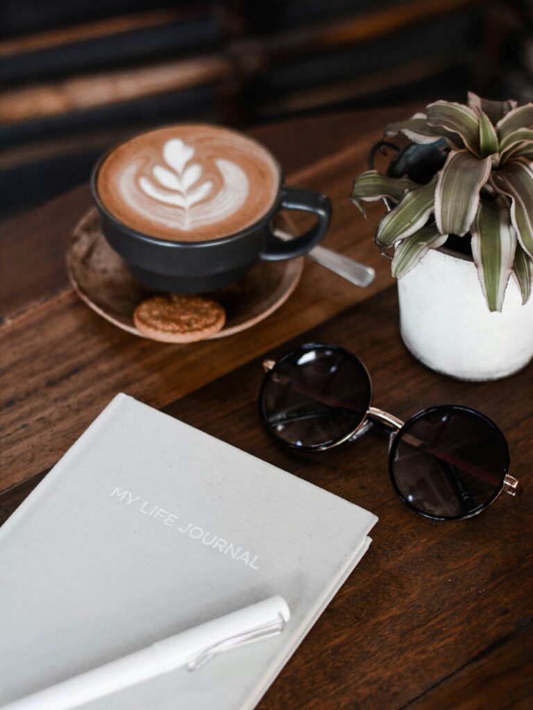 Cappuccino, journal, sunglasses, and plant on a coffee shop table-morning ritual for reflection and goal setting